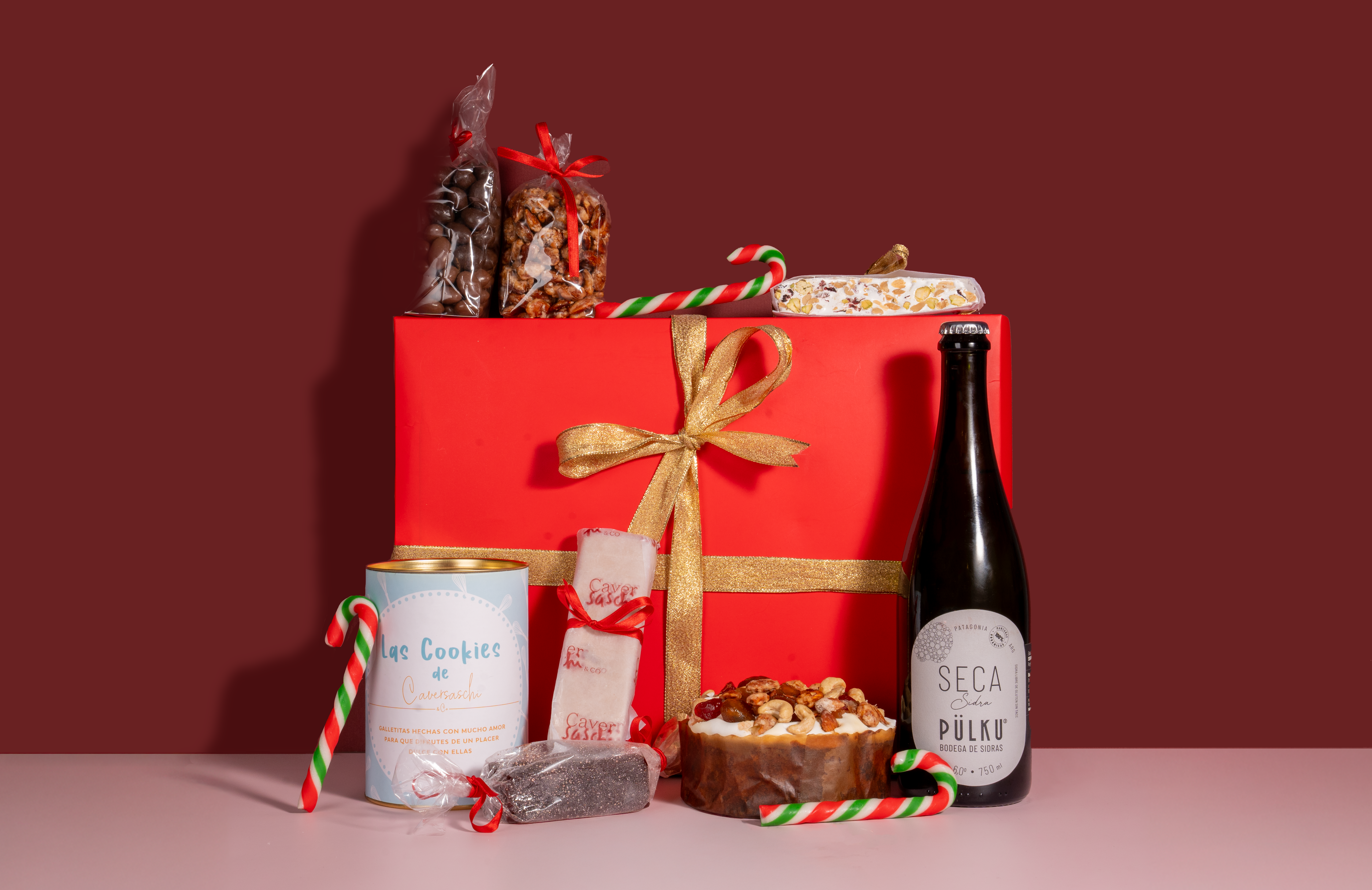 Caja navideña roja con moño dorado rodeada de pan dulce, turrones, sidra y bastones de caramelo
