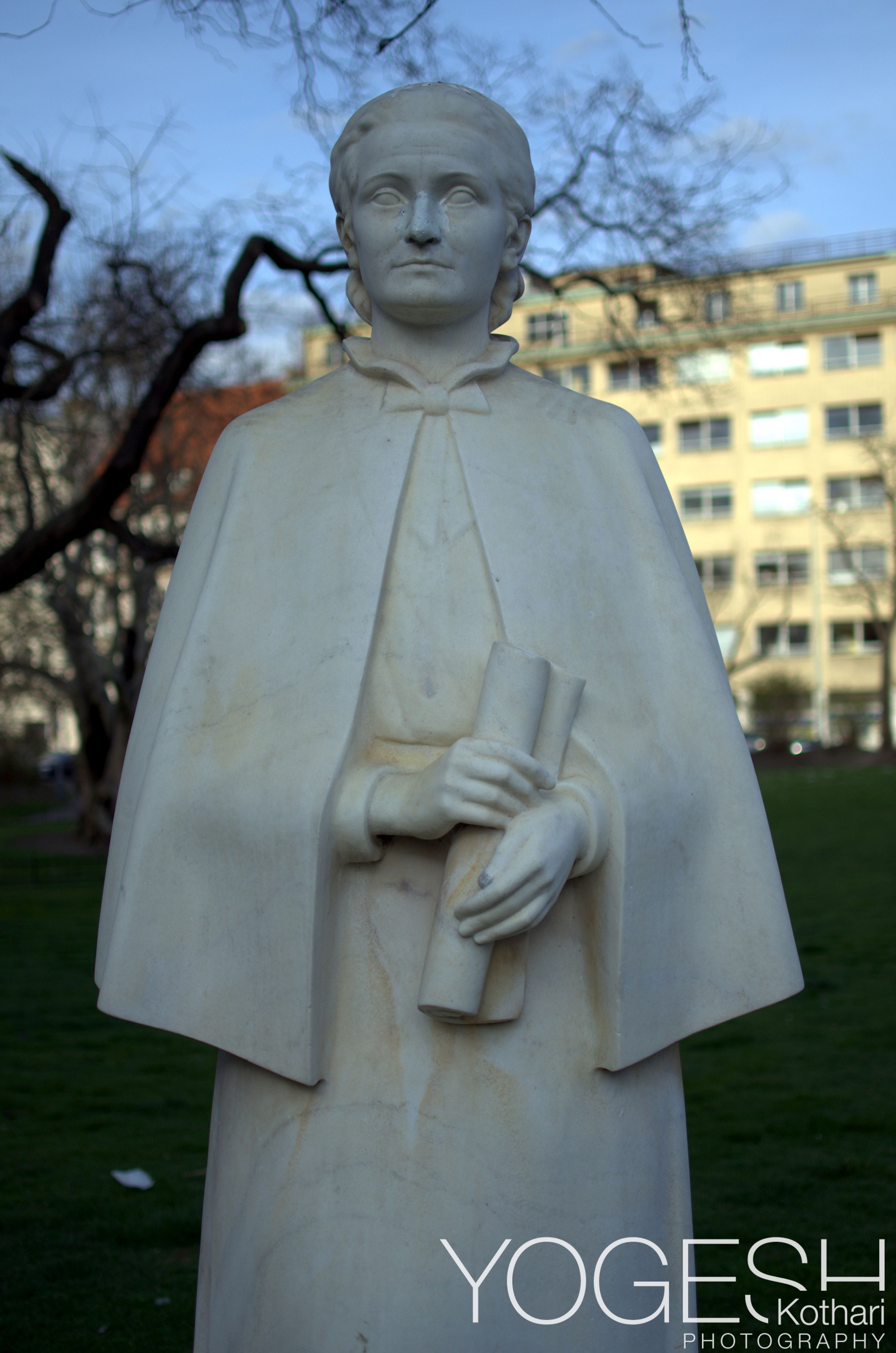 White marble statue of a robed figure holding a scroll, in a park