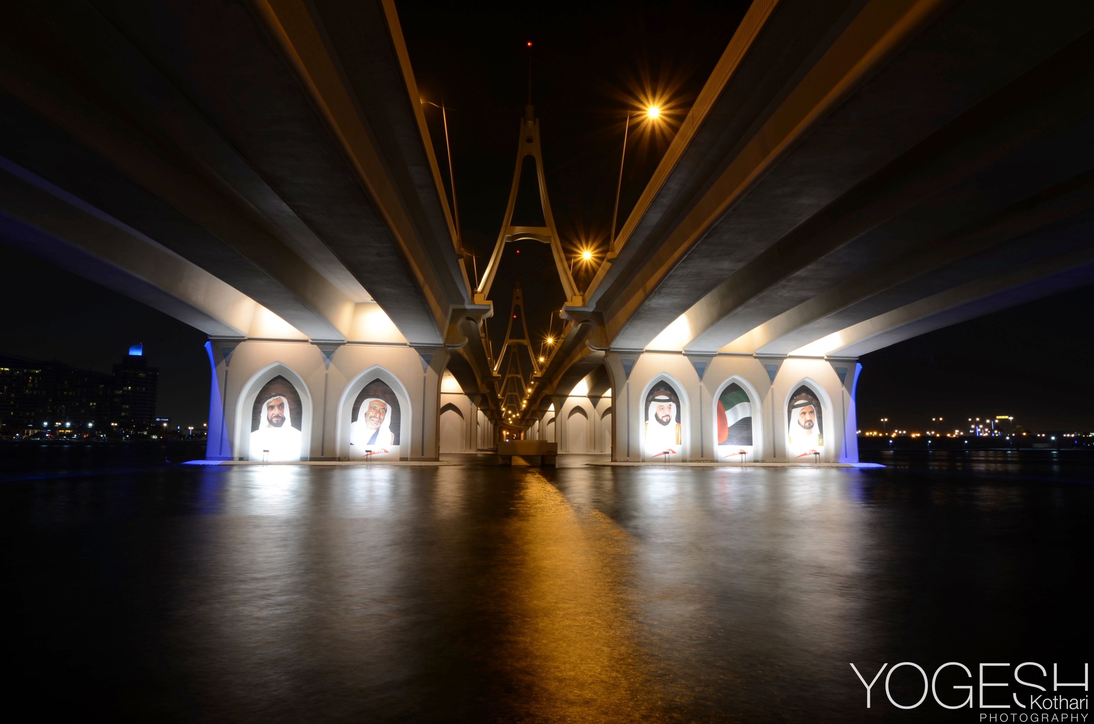Underside of a lit road bridge, its illuminated arches reflected in the water at night