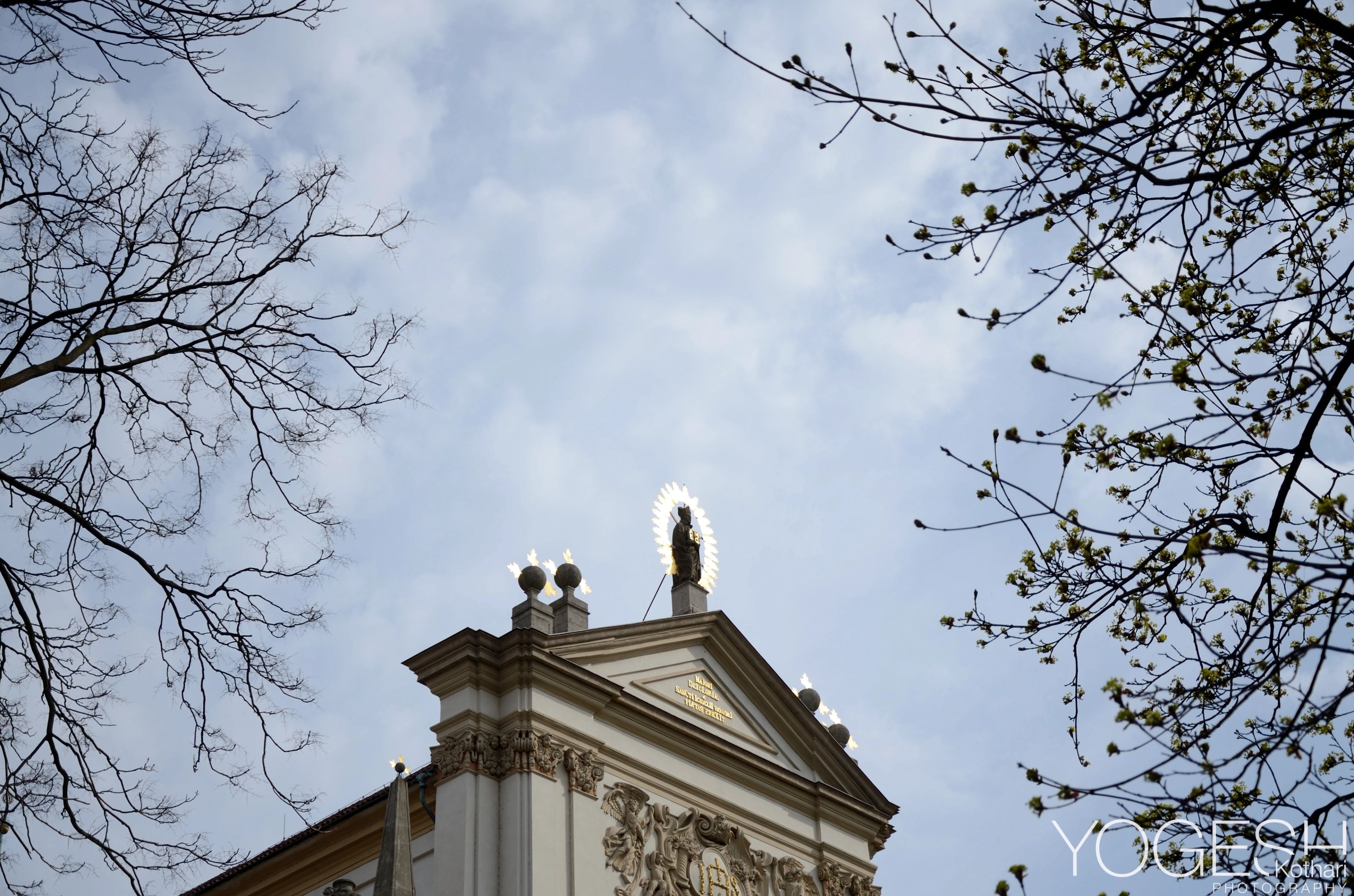 Classical church pediment topped by a statue, framed by bare branches and a cloudy sky