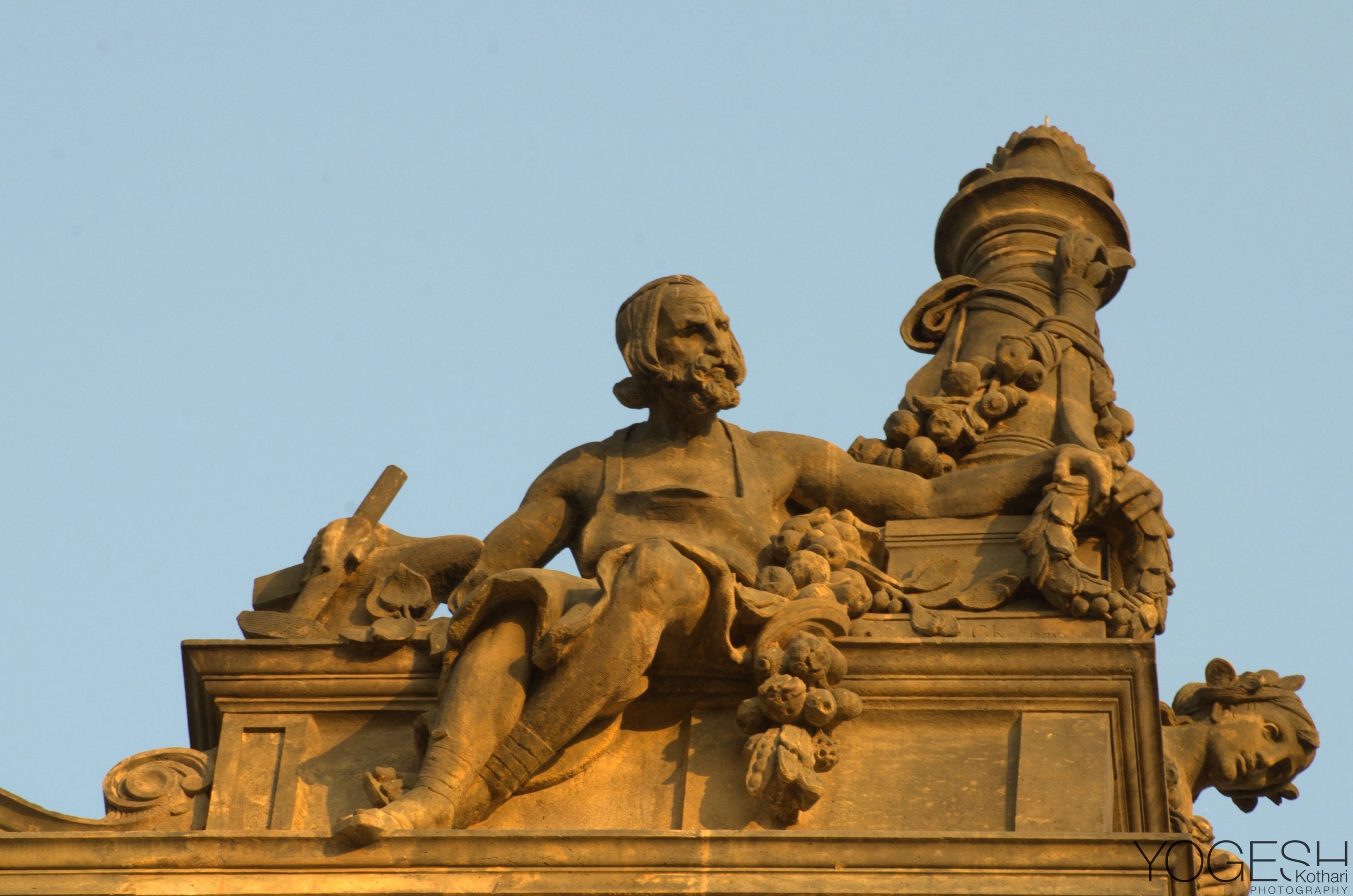Sculpted stone figure reclining beside an urn on a roofline, lit by warm sunlight