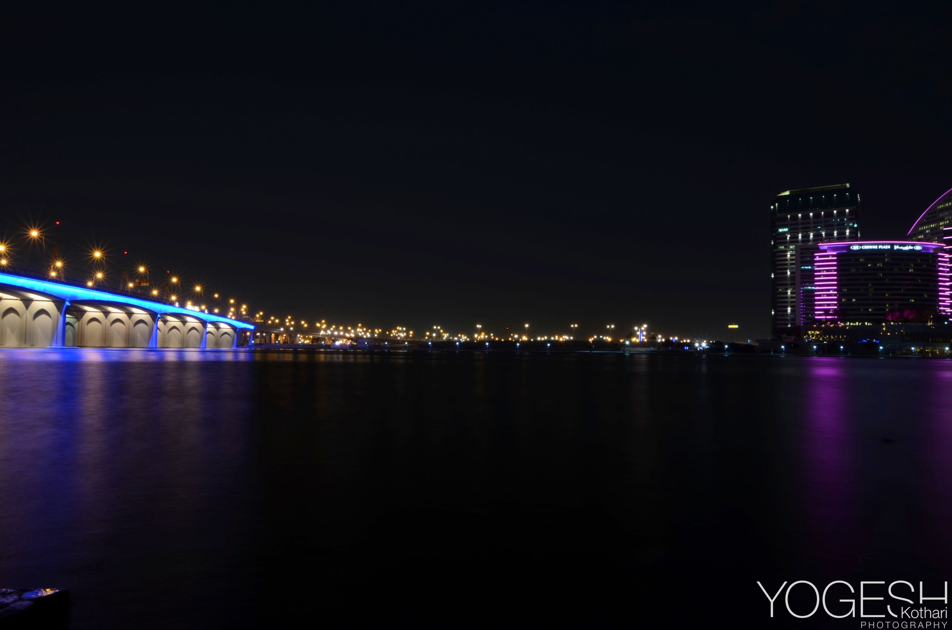 Bridge lit in blue stretching across dark water towards a city skyline at night