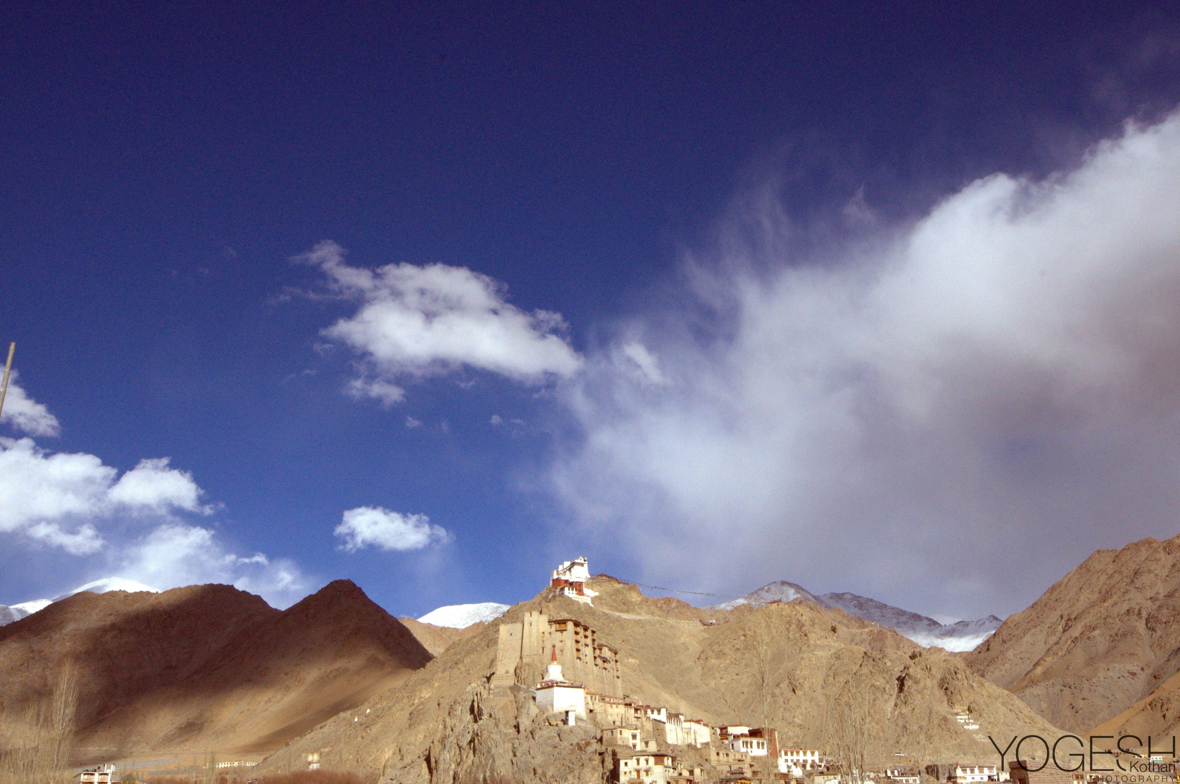 Hilltop monastery above a village, ringed by bare brown mountains under a blue sky with drifting clouds