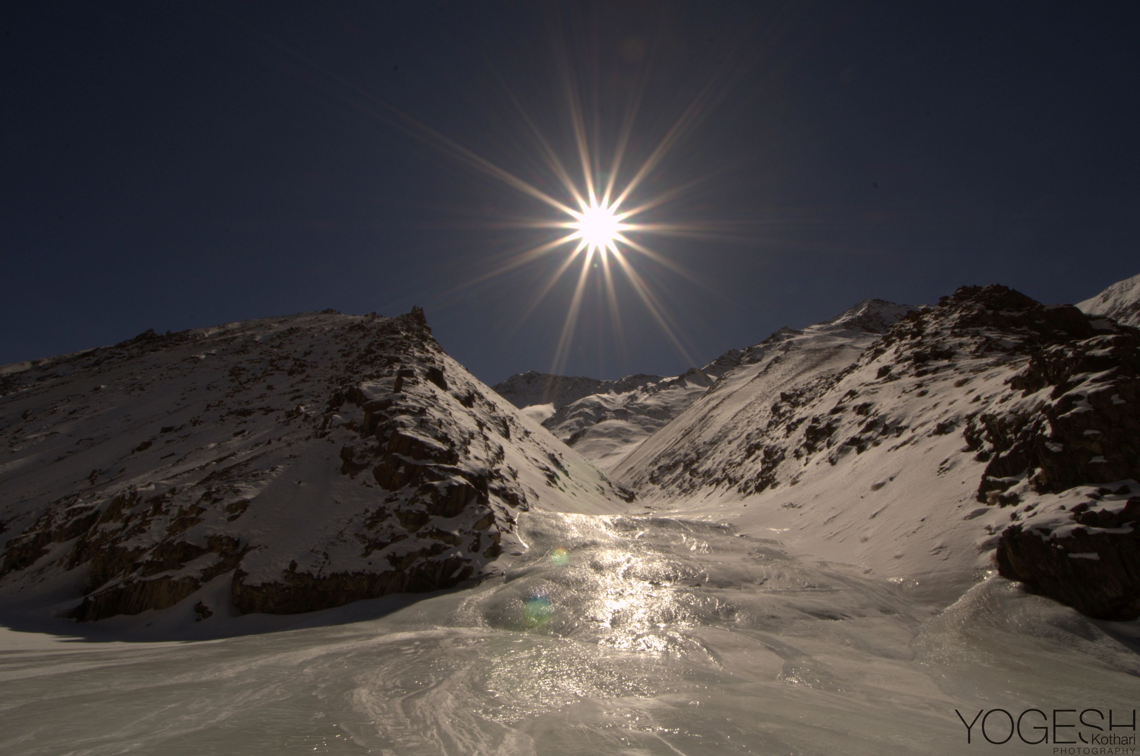 Frozen stream winding between snowy slopes, lit by a bright star-shaped sun