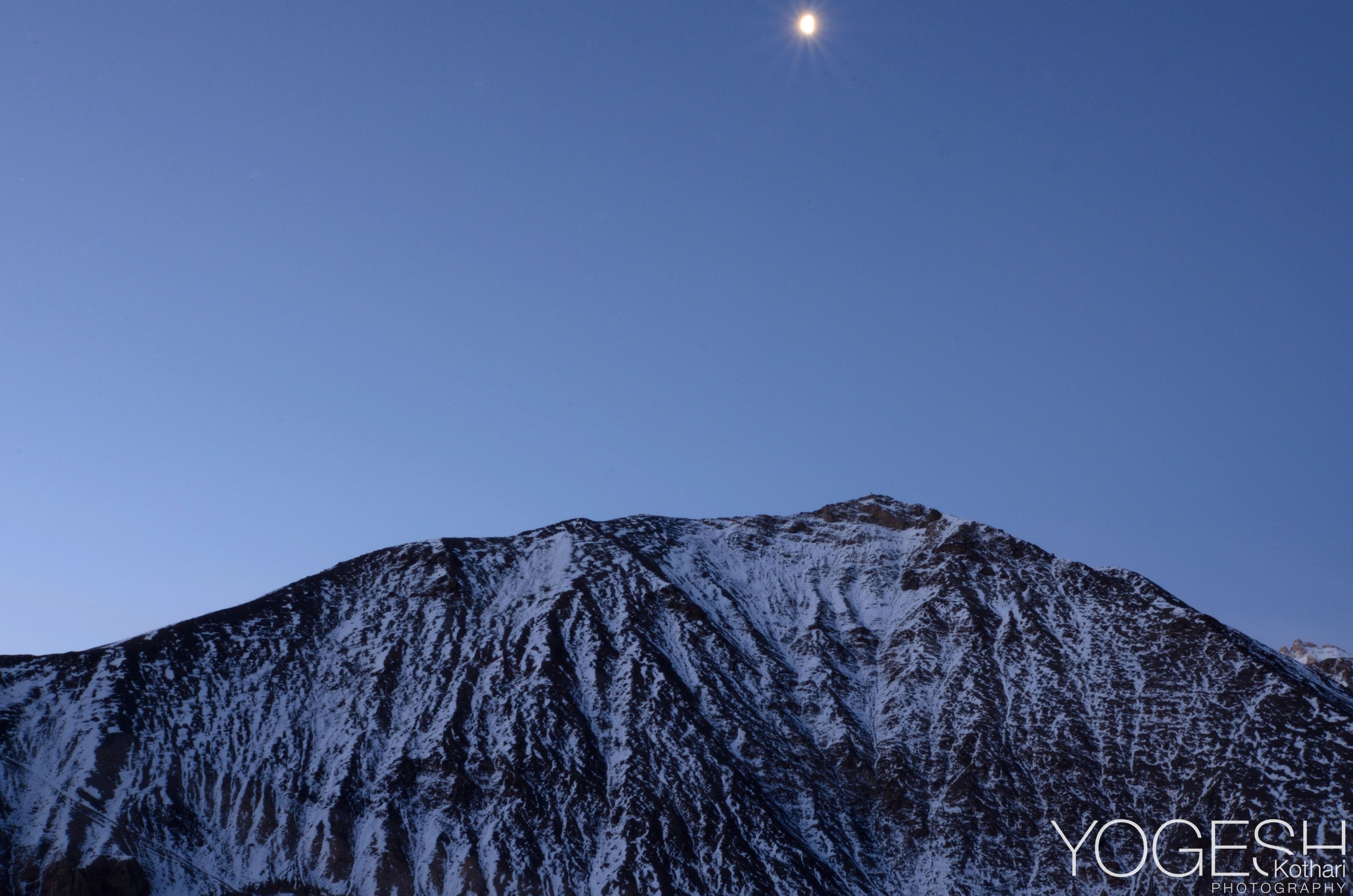Small moon in a clear blue sky above a snow-dusted mountain face