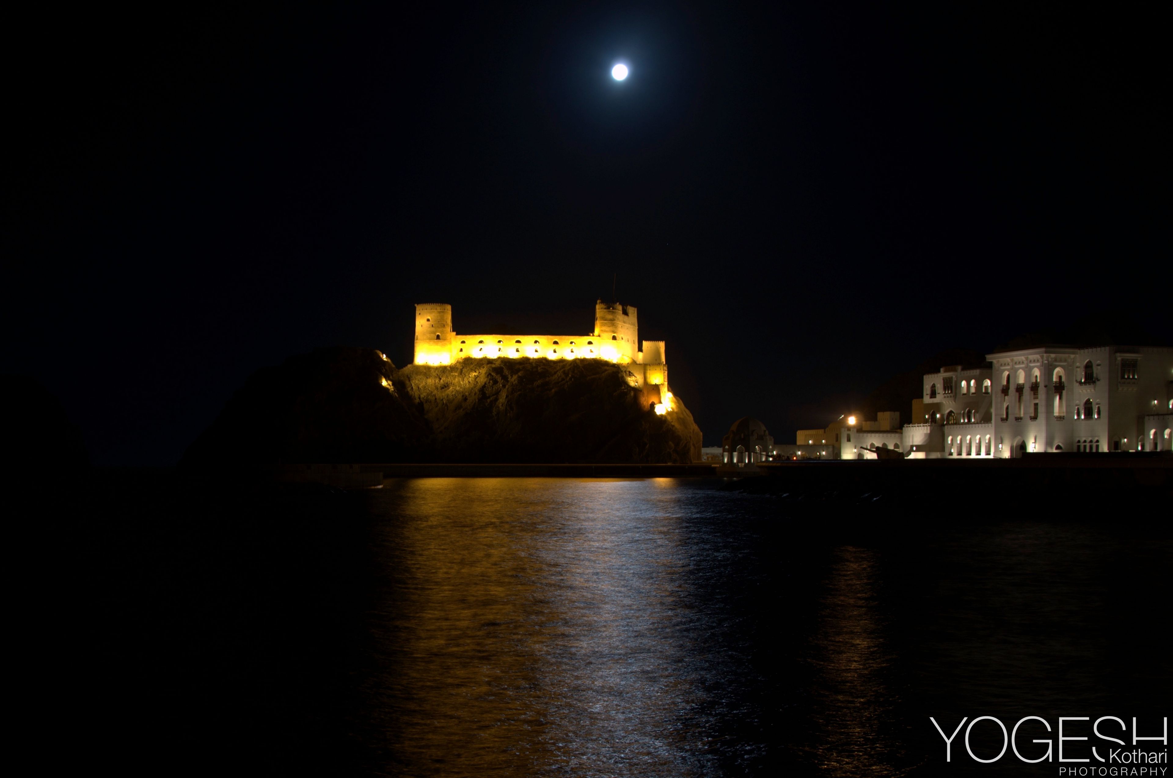 Floodlit hilltop fort under a full moon, its lights reflected in the water below