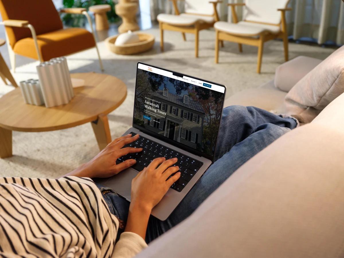 Person on a sofa using a laptop showing the Savannah Walks homepage hero, Savannah Walking Tours, over a photo of a historic Savannah home