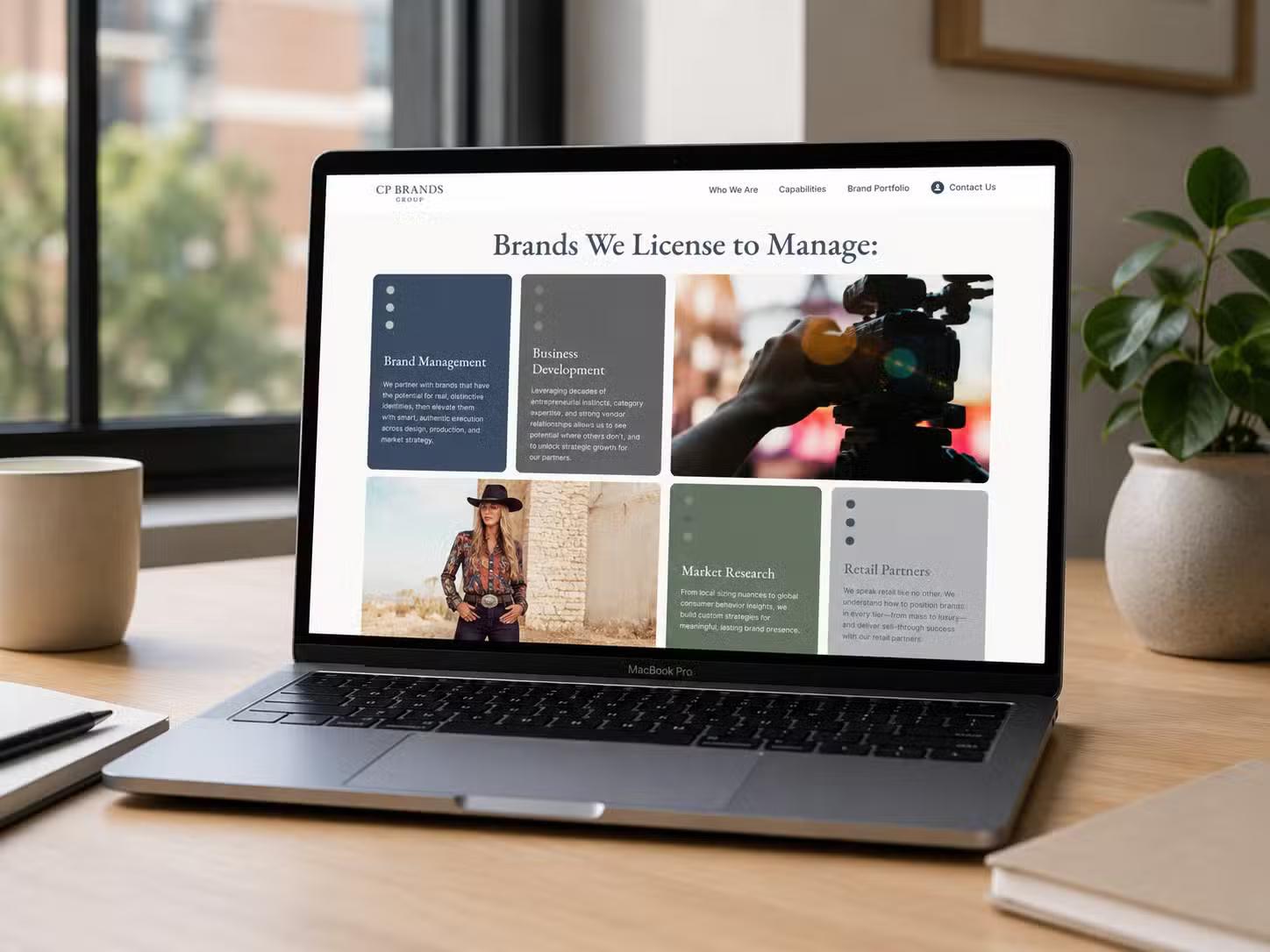 Laptop on a wooden desk near a window displaying the CP Brands Group website "Brands We License to Manage" page, a grid of feature blocks paired with lifestyle photography.