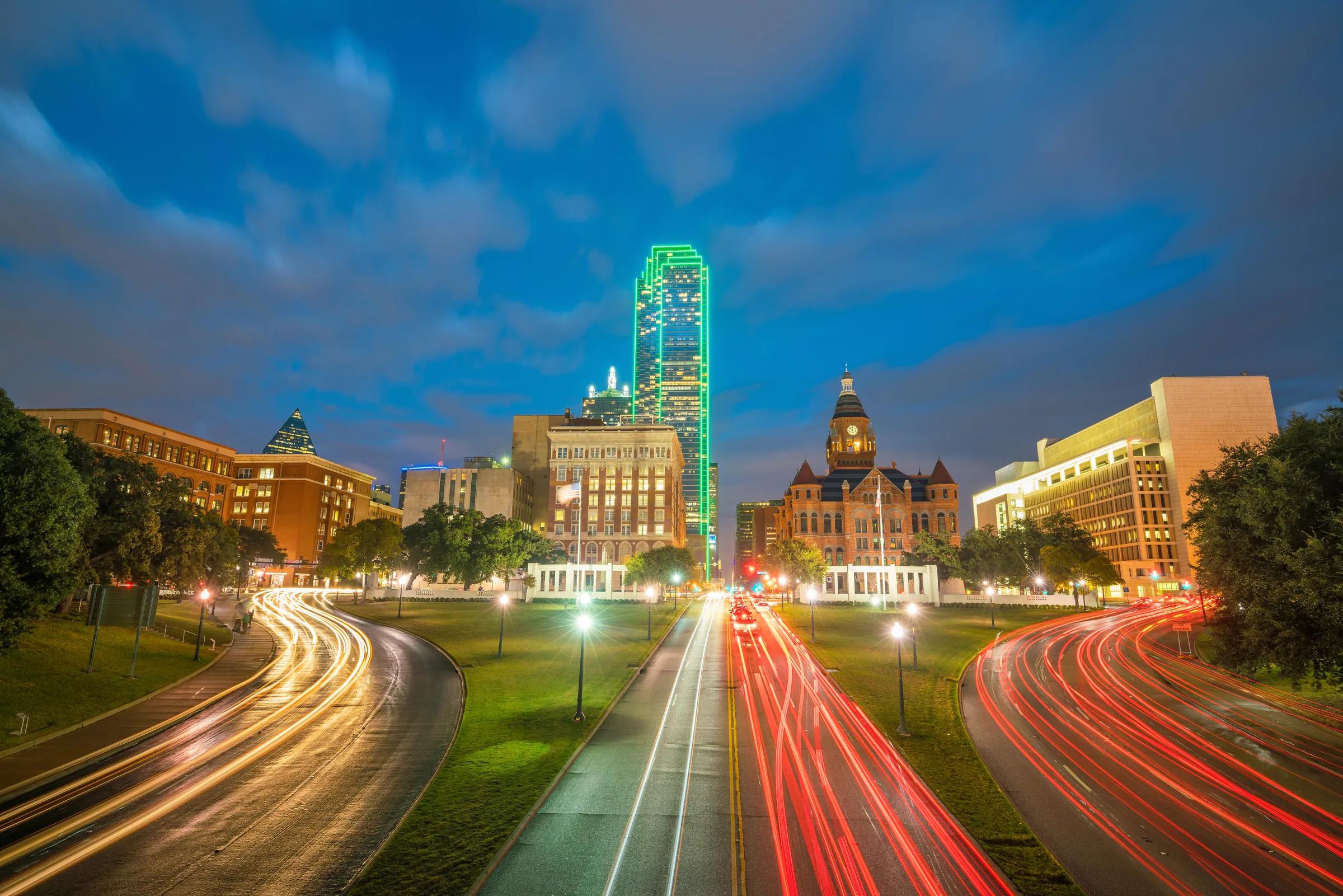 Light trails leading toward the Downtown Dallas skyline under a blue evening sky