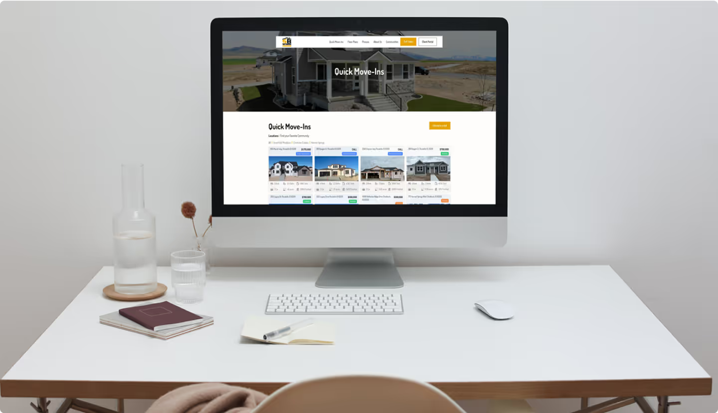 iMac on clean white desk showing Satterfield Builders Quick Move-Ins page with panoramic community hero photo and grid of available property listing cards with home exteriors and details