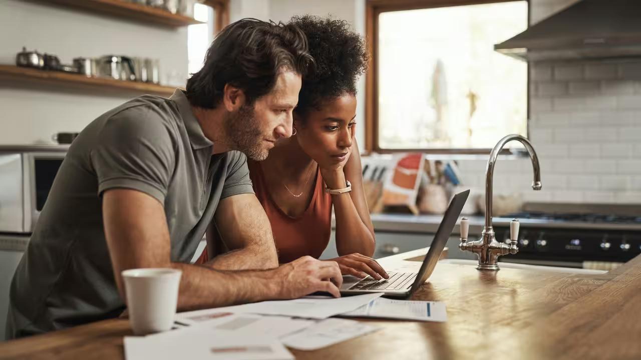 Two homeowners at a kitchen table researching contractors on a laptop with project paperwork spread out in front of them.