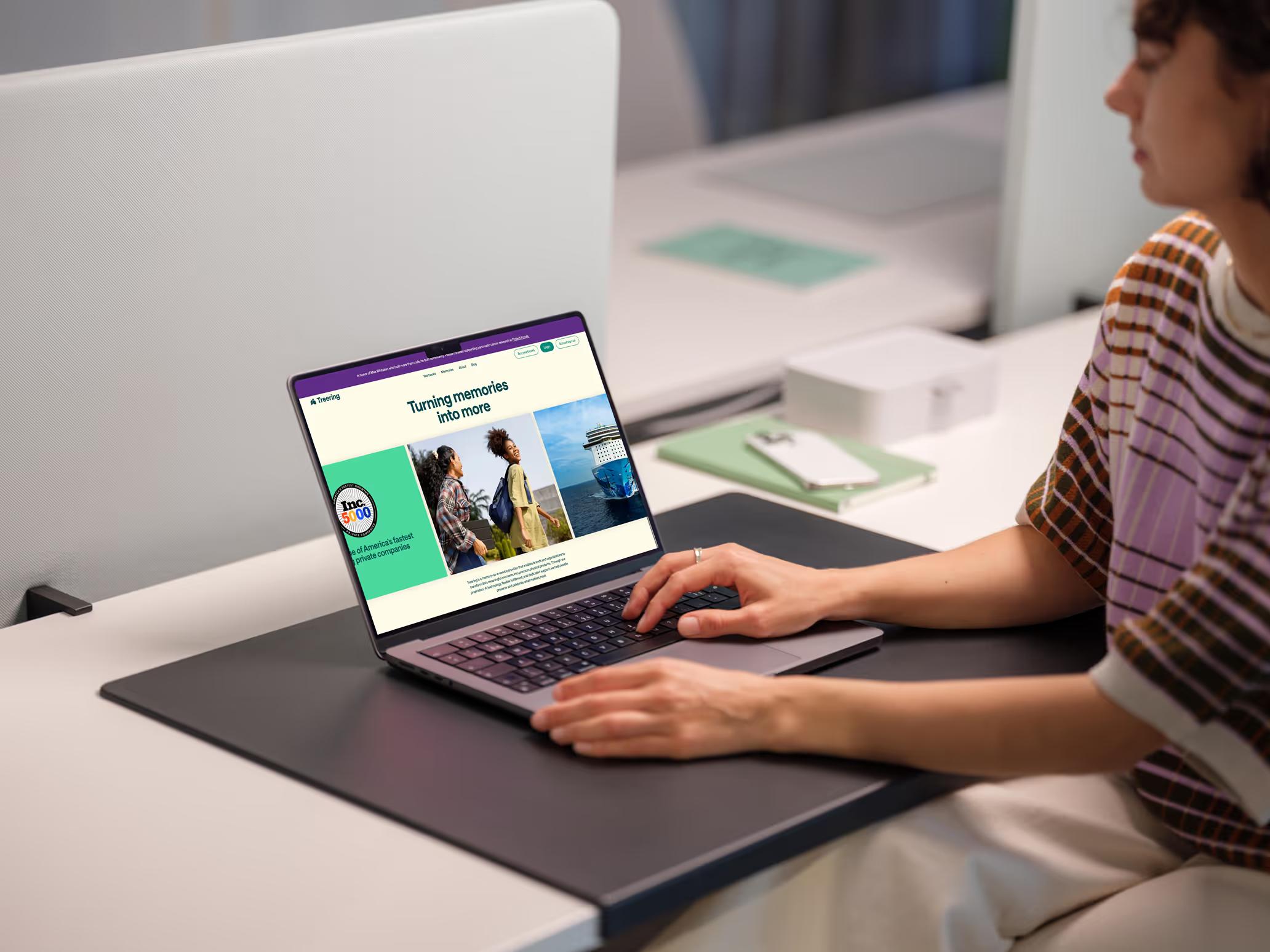 Woman at office desk browsing TreeRing homepage on MacBook showing Turning memories into more hero with Inc 500 badge and colorful lifestyle imagery