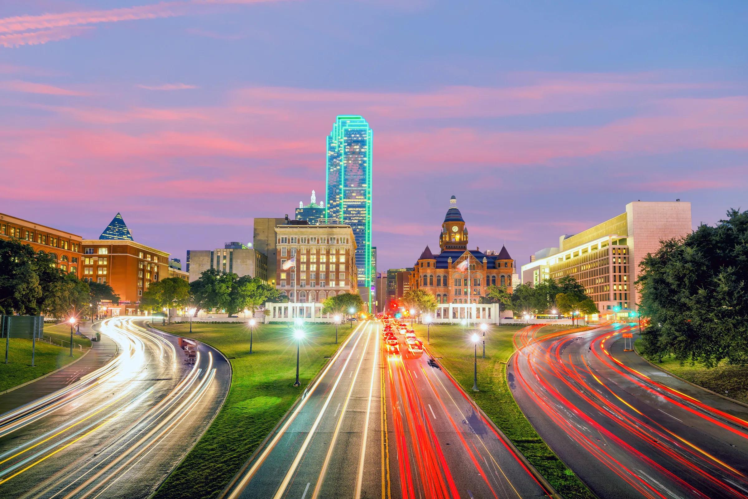 Light trails leading toward the Downtown Dallas skyline at dusk under a pink sky