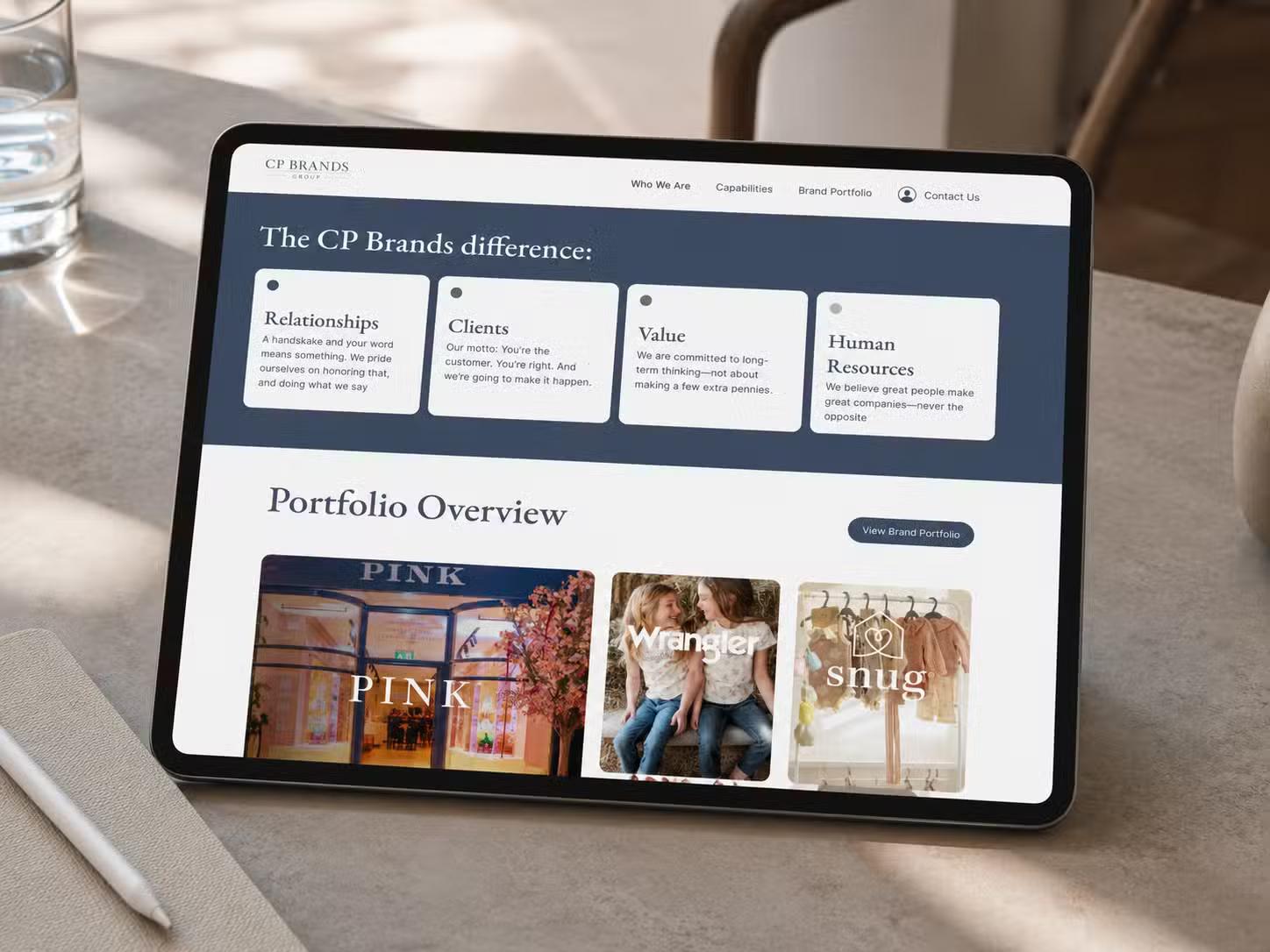 Tablet on a stone surface showing the CP Brands Group website, with a "The CP Brands difference" section and a Portfolio Overview grid featuring Pink, Wrangler, and Snug brand cards.