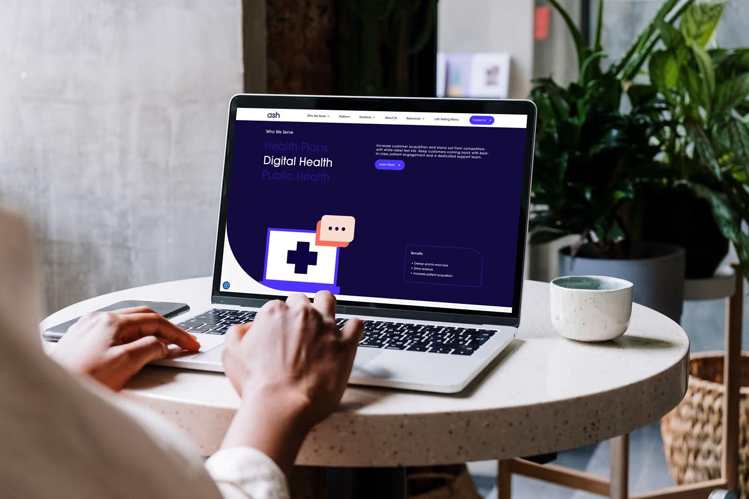 MacBook on round café table showing Ash Wellness Who We Serve page with Health Plans, Digital Health, and Public Health sections on dark navy background