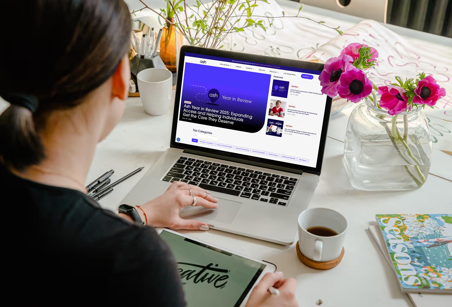 Woman at creative desk browsing Ash Wellness blog page on MacBook with Year in Review featured article, pink flowers in vase, and coffee cup