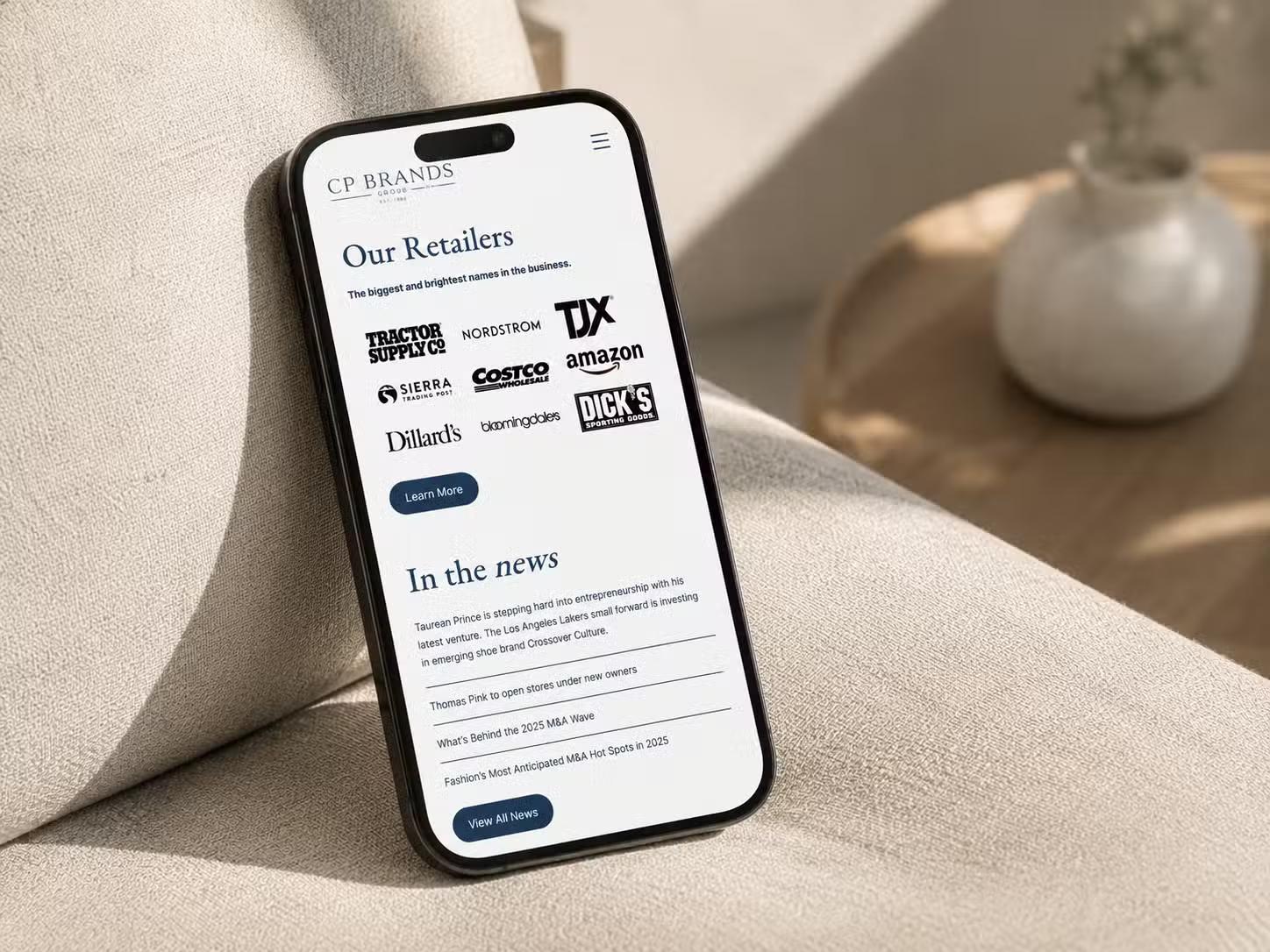 iPhone propped on a couch showing the CP Brands Group mobile website, with an "Our Retailers" section of logos including Tractor Supply, Nordstrom, TJX, Costco, Amazon, and Dillard's, followed by an "In the news" list.