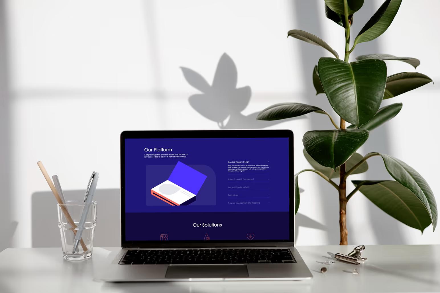 MacBook on white desk next to plant showing Ash Wellness Our Platform page with dark navy section featuring 3D book illustration and Branded Program Design description
