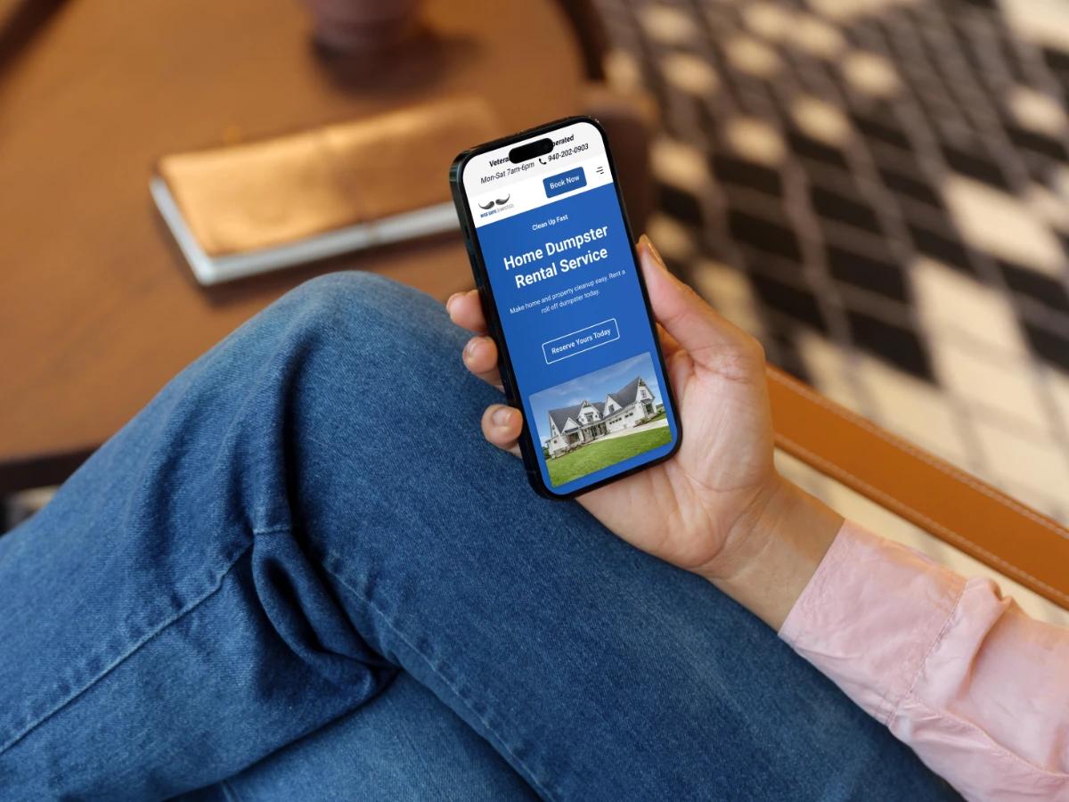 Hand holding a phone showing the Wise Guys Dumpsters mobile Home Dumpster Rental Service page with a Reserve Yours Today button