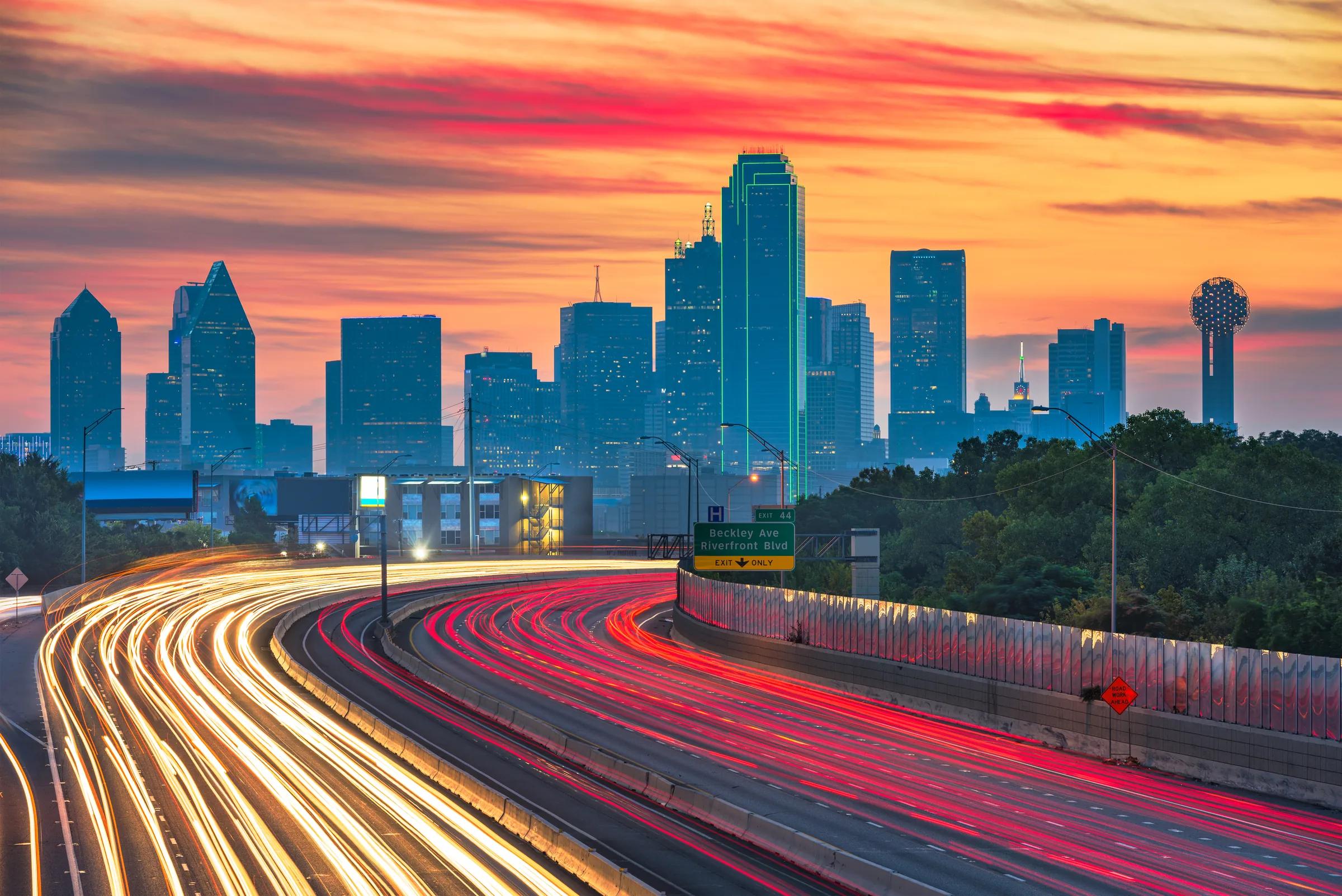 Vehicle light trails on a Dallas highway with the skyline at sunset