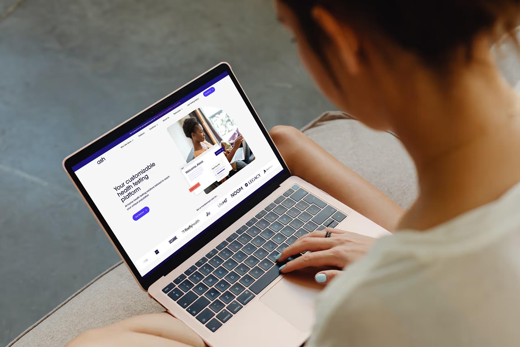 Woman on couch browsing Ash Wellness homepage on MacBook showing Your customizable health testing platform headline with hero image and partner logos