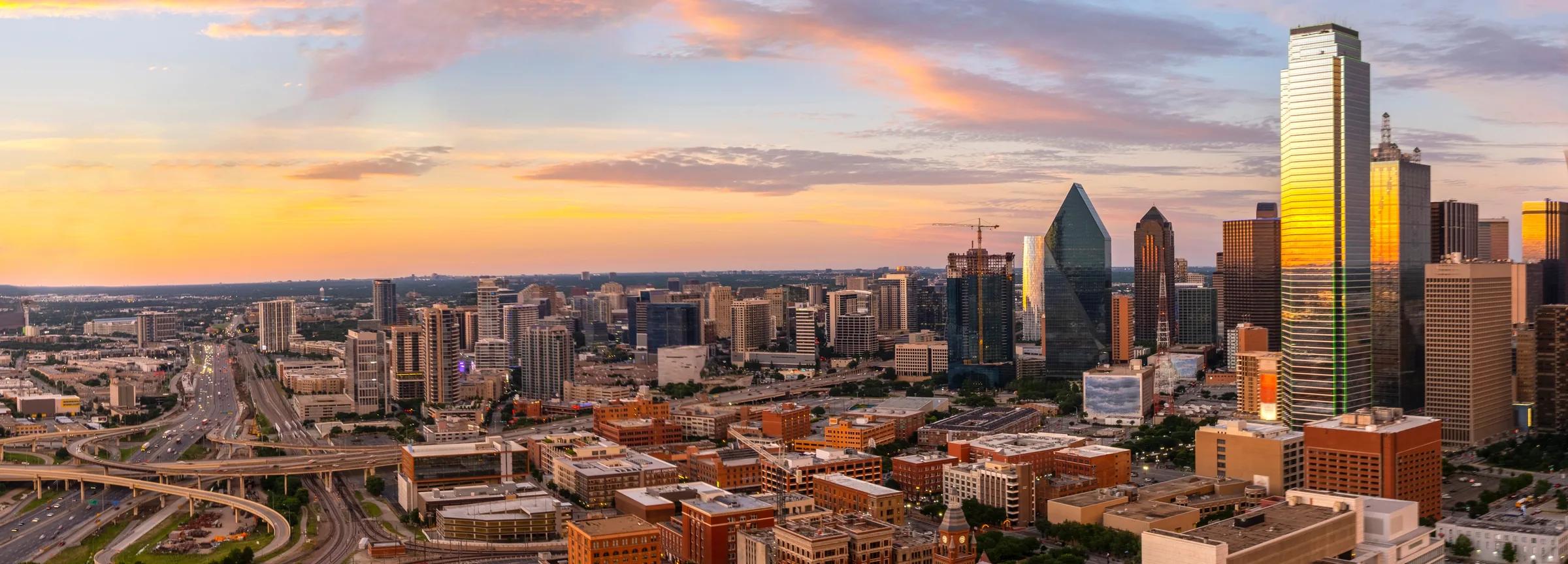 The Dallas skyline at sunrise under a pastel sky, with a highway in the foreground