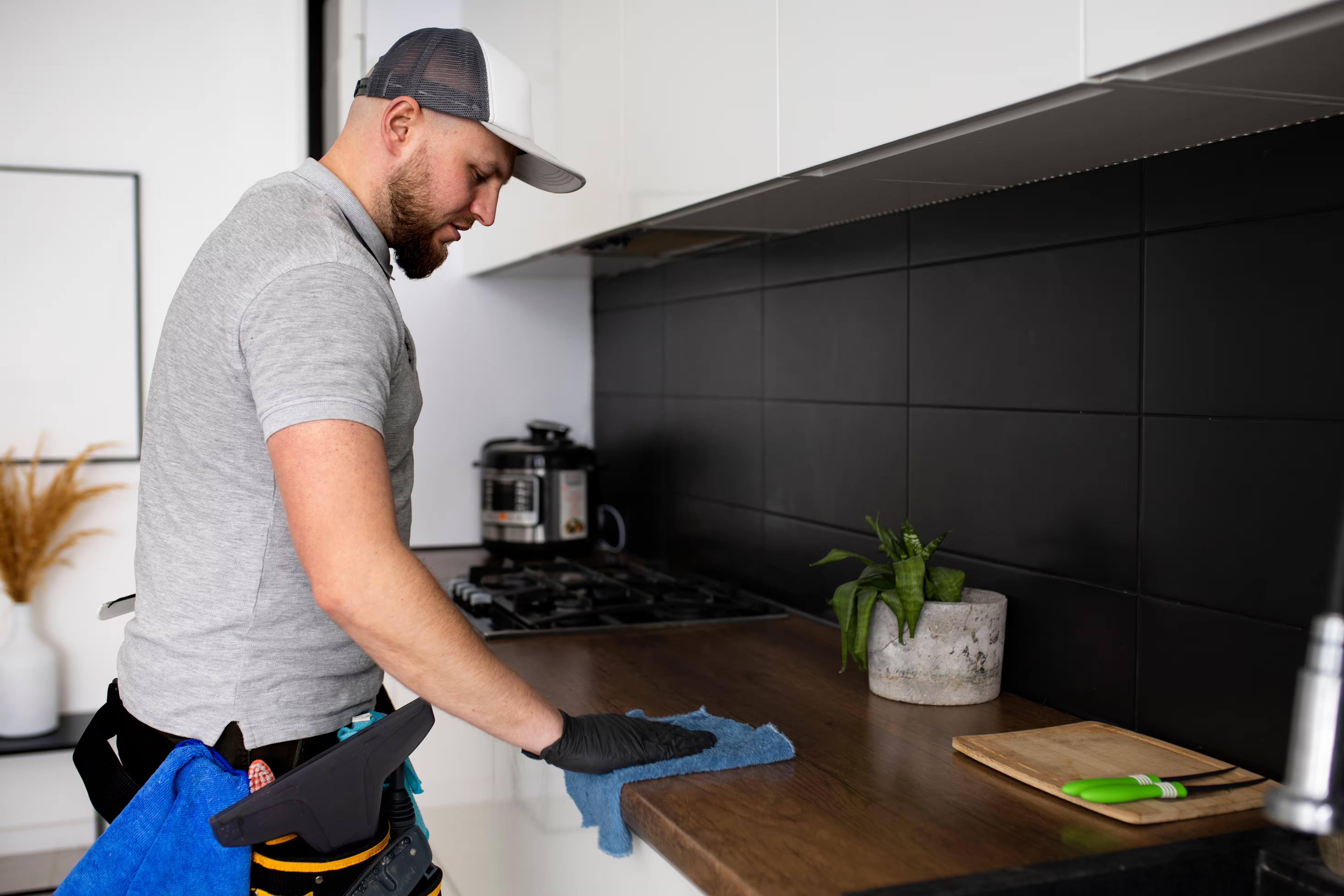 A professional cleaner in uniform wiping down a kitchen counter with a tool belt of supplies, doing the specific job a cleaning company's homepage should name.