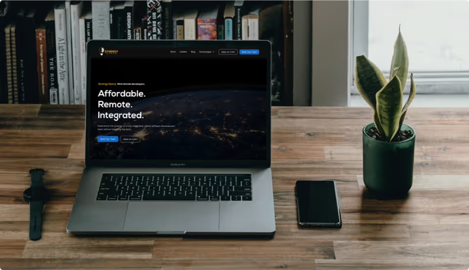 MacBook on wooden desk showing Synergy Sparq homepage with Affordable Remote Integrated headline over dark globe hero image, next to plant and phone