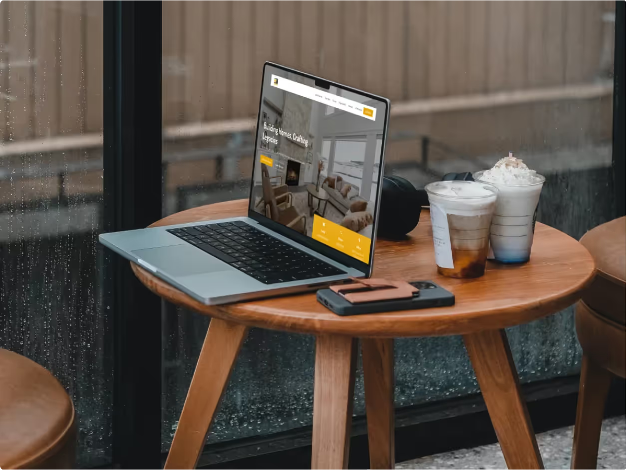 MacBook at café table with iced coffees showing Satterfield Builders homepage hero Building Homes Crafting Legacies with warm living room interior