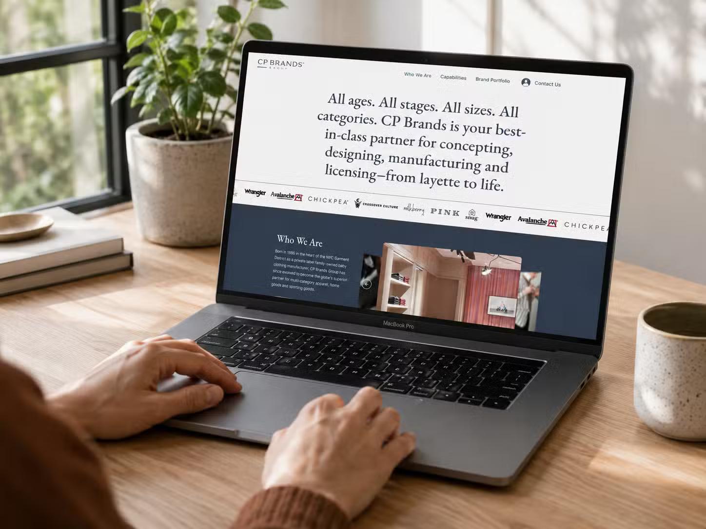 Laptop on a wooden desk displaying the CP Brands Group website homepage, with a hero headline reading "All ages. All stages. All sizes. All categories." above a row of brand logos; a person's hands rest on the keyboard.