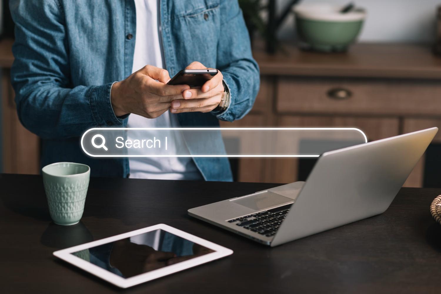 man searching on his phone with a computer and tablet presence along with a search bar