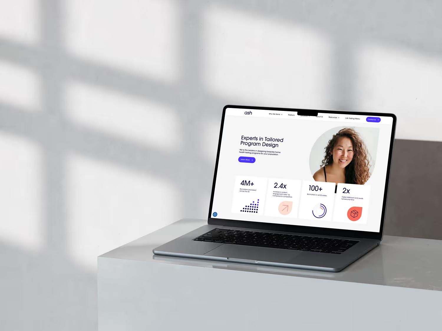 MacBook on minimal white shelf displaying Ash Wellness Experts in Tailored Program Design page with woman portrait and stat cards showing 4M+, 2.4x, 100+, and 2x metrics