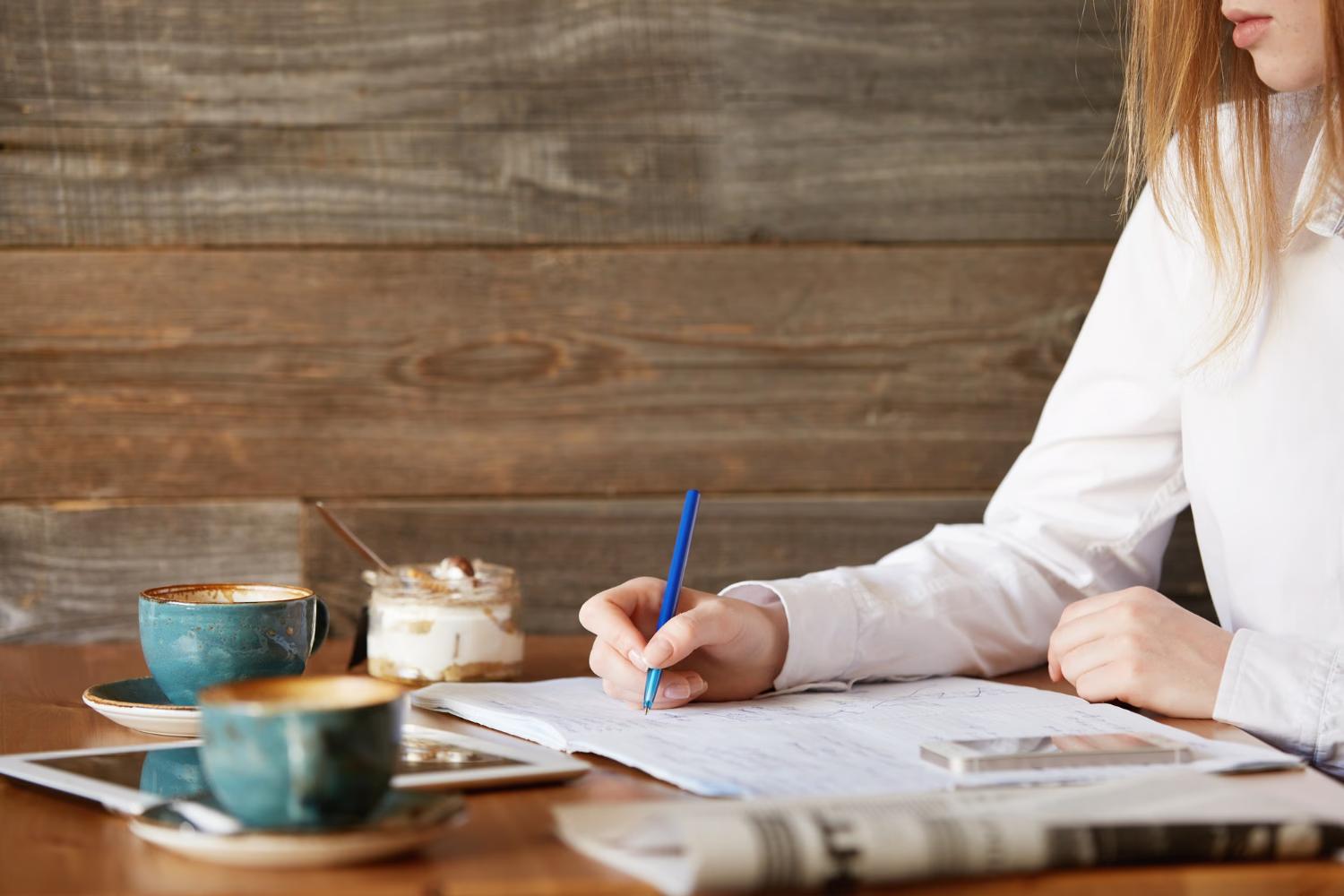 woman at a table with pen and paper along with coffee and dessert doing keyword brainstorming