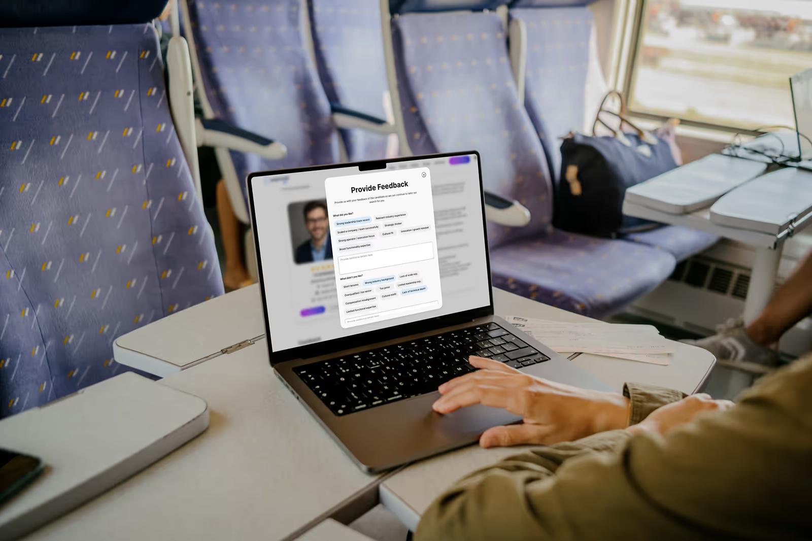 MacBook on train seat tray table showing PopCandi AI Provide Feedback form for a candidate with rating fields and text inputs