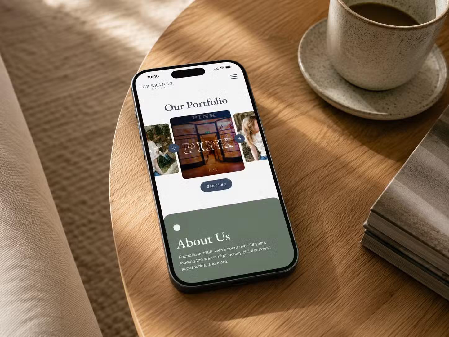 iPhone on a wooden table beside a coffee cup showing the CP Brands Group mobile website, with an "Our Portfolio" carousel and a green "About Us" section below.