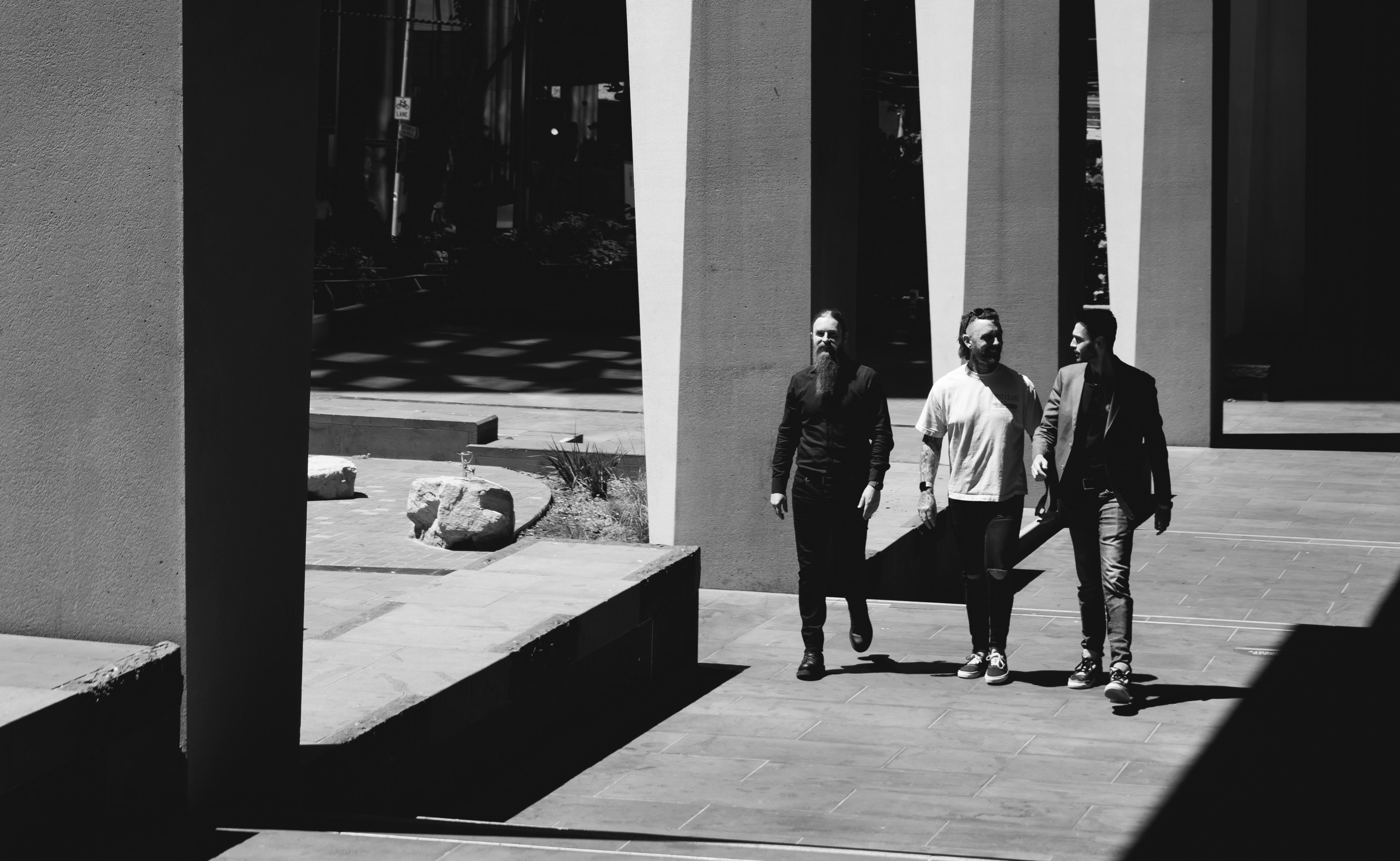 This black-and-white photograph captures three people walking confidently side by side through a dramatically lit urban space. The bold, architectural columns create stark shadows and contrast, enhancing the artistic impact of the image. Each individual walks with purpose, suggesting determination, unity, and collaboration. The interplay between light and shadow emphasises their movement forward, symbolising progress and teamwork against an architecturally striking backdrop. The overall mood is sophisticated and cinematic, capturing an everyday moment elevated through thoughtful composition and lighting.