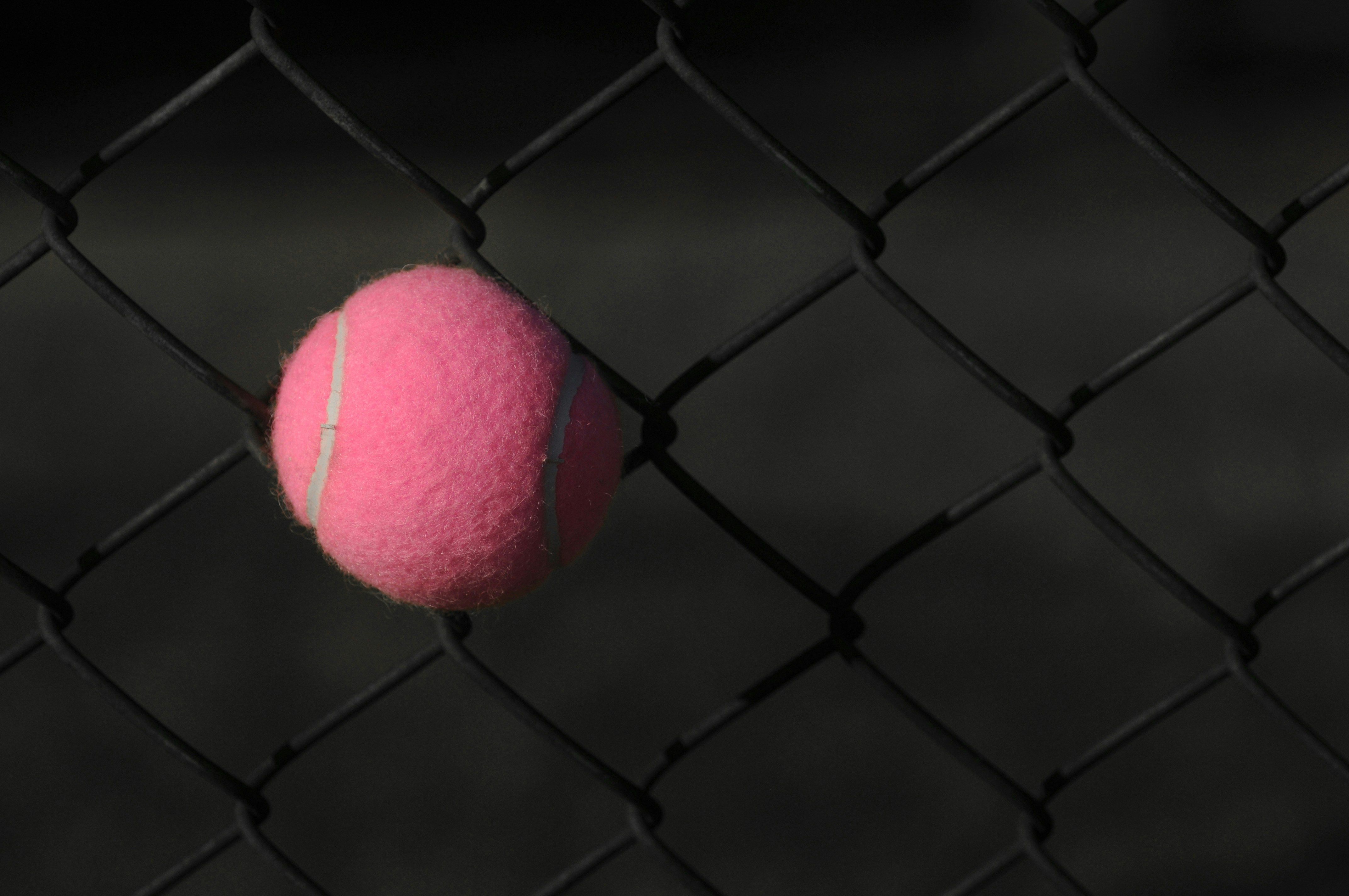 A pink tennis ball caught in a chain link fence.