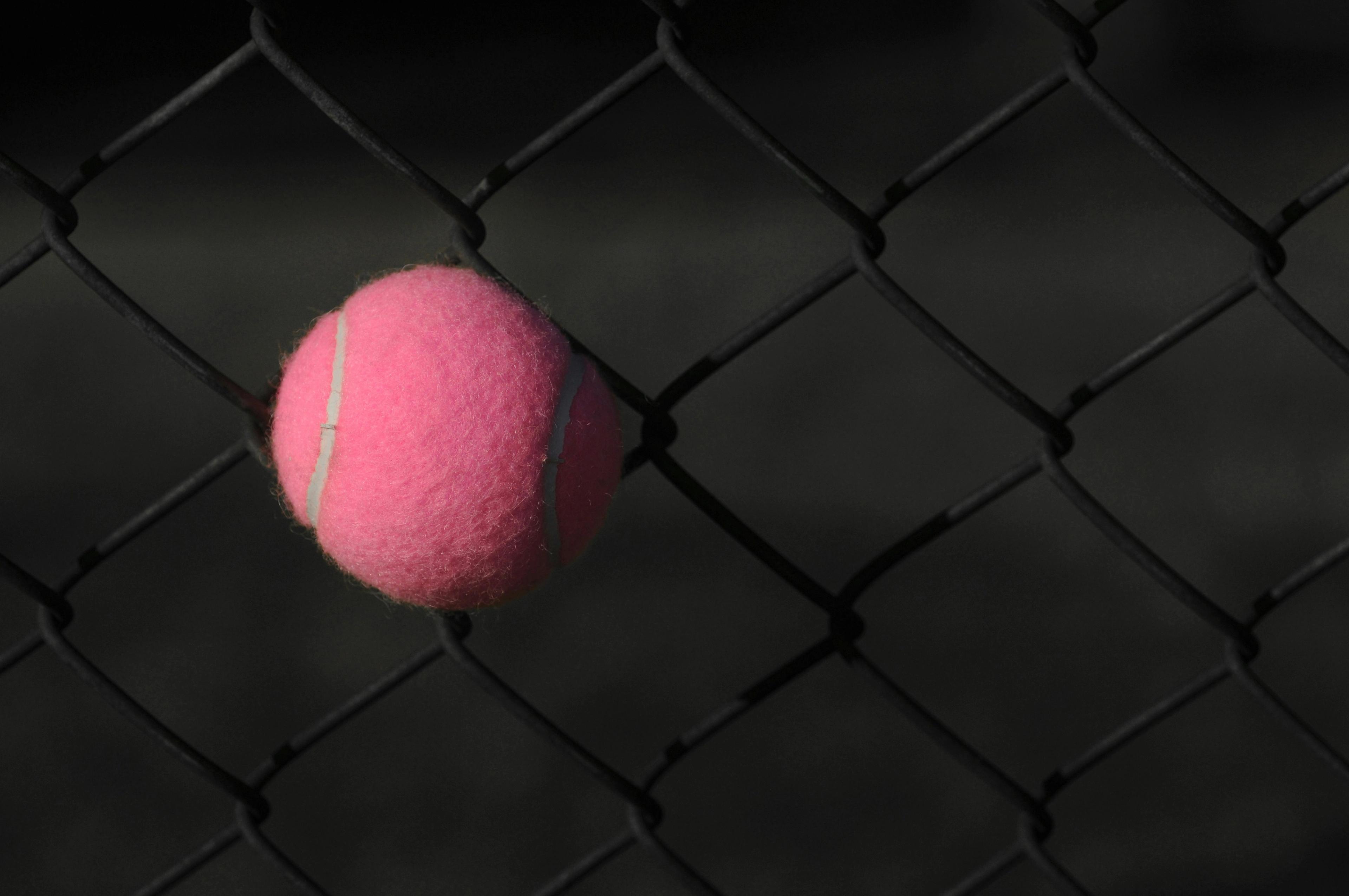A pink tennis ball caught in a chain link fence.
