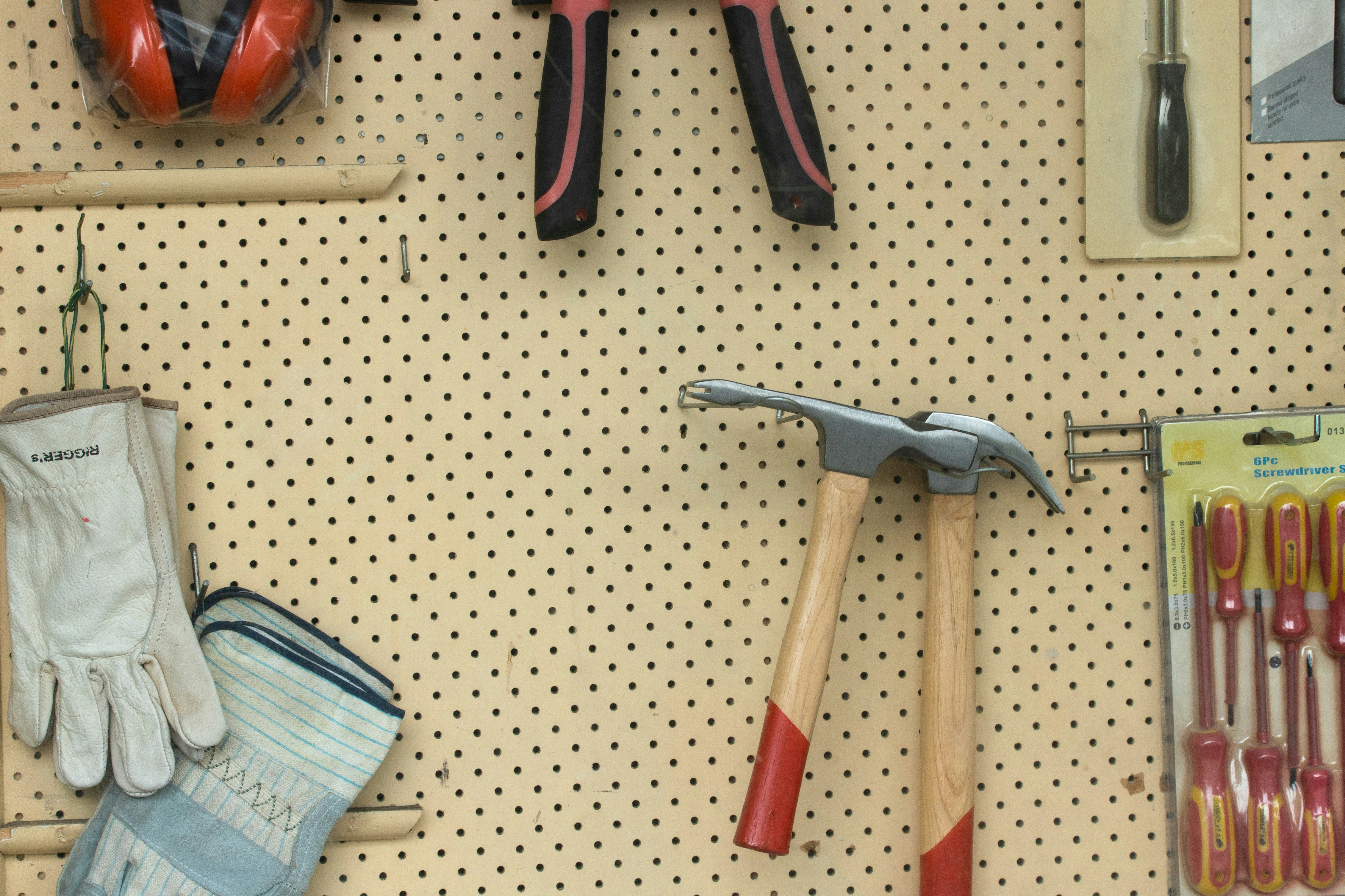 a hammer , pliers , gloves , and screwdrivers are hanging on a peg board .
