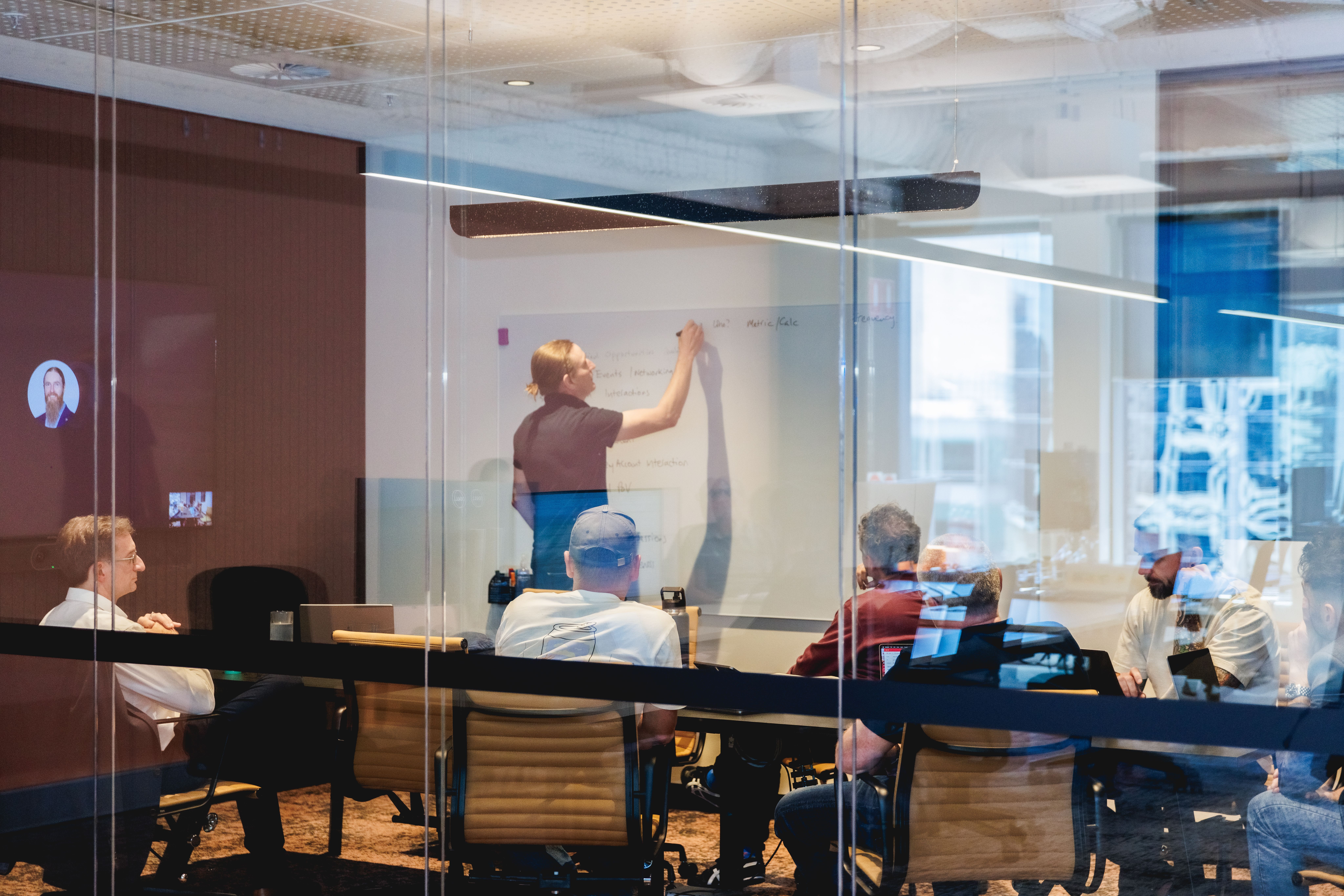 This dynamic image captures a lively team discussion within a modern conference room viewed through transparent glass walls. A person is actively writing on a whiteboard, suggesting brainstorming or strategic planning is underway. Team members seated around the table are focused attentively on the presenter, indicative of active engagement and participation. A virtual participant is visible on the large screen, highlighting remote collaboration. The reflections and lighting enhance the contemporary feel of the space, suggesting a professional environment that encourages creativity, innovation, and teamwork.