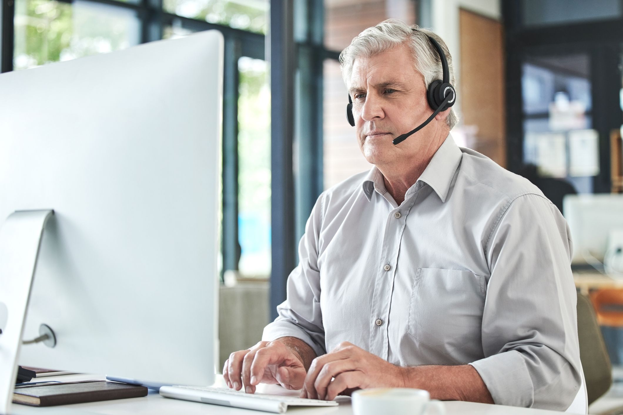 An older man uses a computer at work wearing a headset.