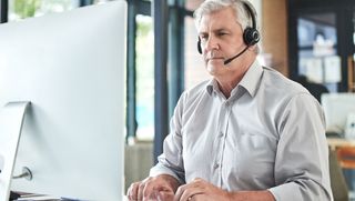An older man uses a computer at work wearing a headset.