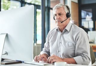 An older man uses a computer at work wearing a headset.