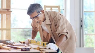 A older man with safety glasses working in a carpentry shop, measuring a table