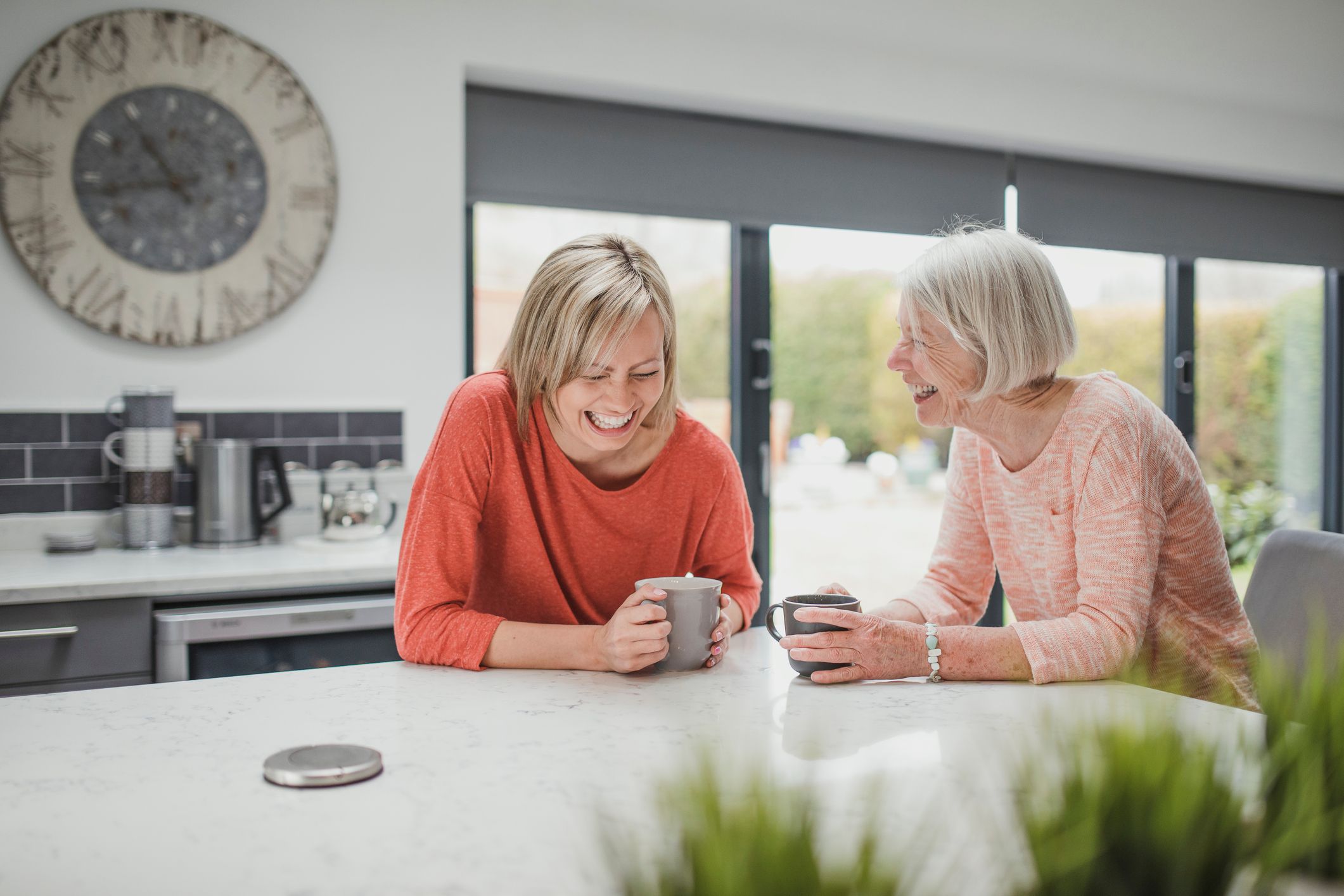 Two women sitting at a kitchen island, laughing with each other with mugs in hand.