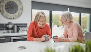 Two women sitting at a kitchen island, laughing with each other with mugs in hand.