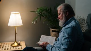 Older person seated in dark room reading book by lamp light