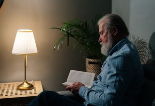 Older person seated in dark room reading book by lamp light