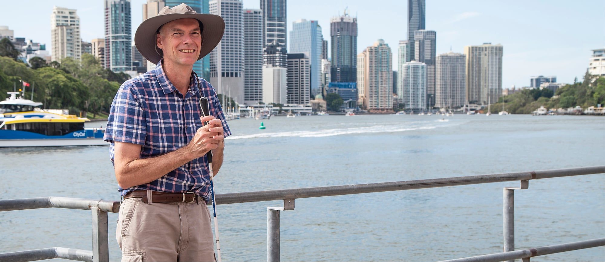 A man holding a white cane stands in front of a lake and smiles at the camera.