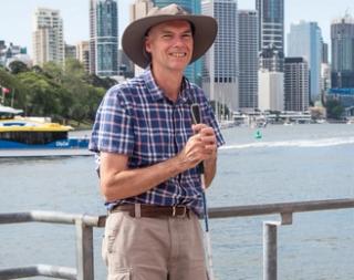 A man holding a white cane stands in front of a lake and smiles at the camera.